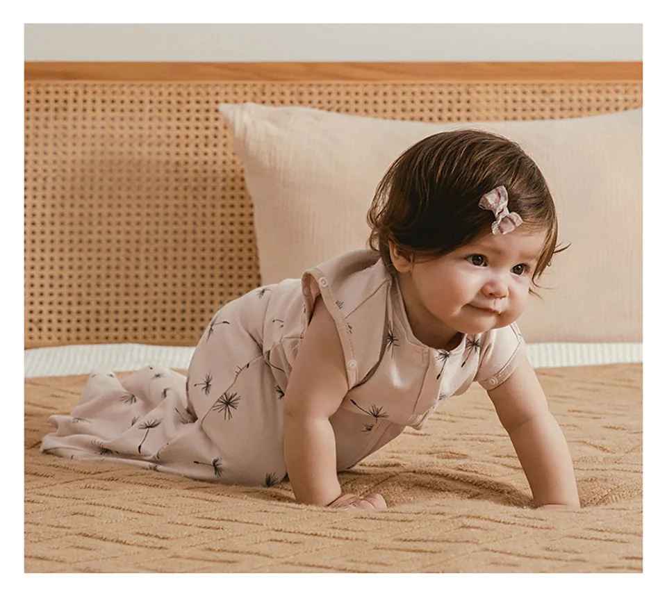 petite fille qui est a quatre pattes sur le lit avec sa gigoteuse écru avec des fleurs de pissenlits